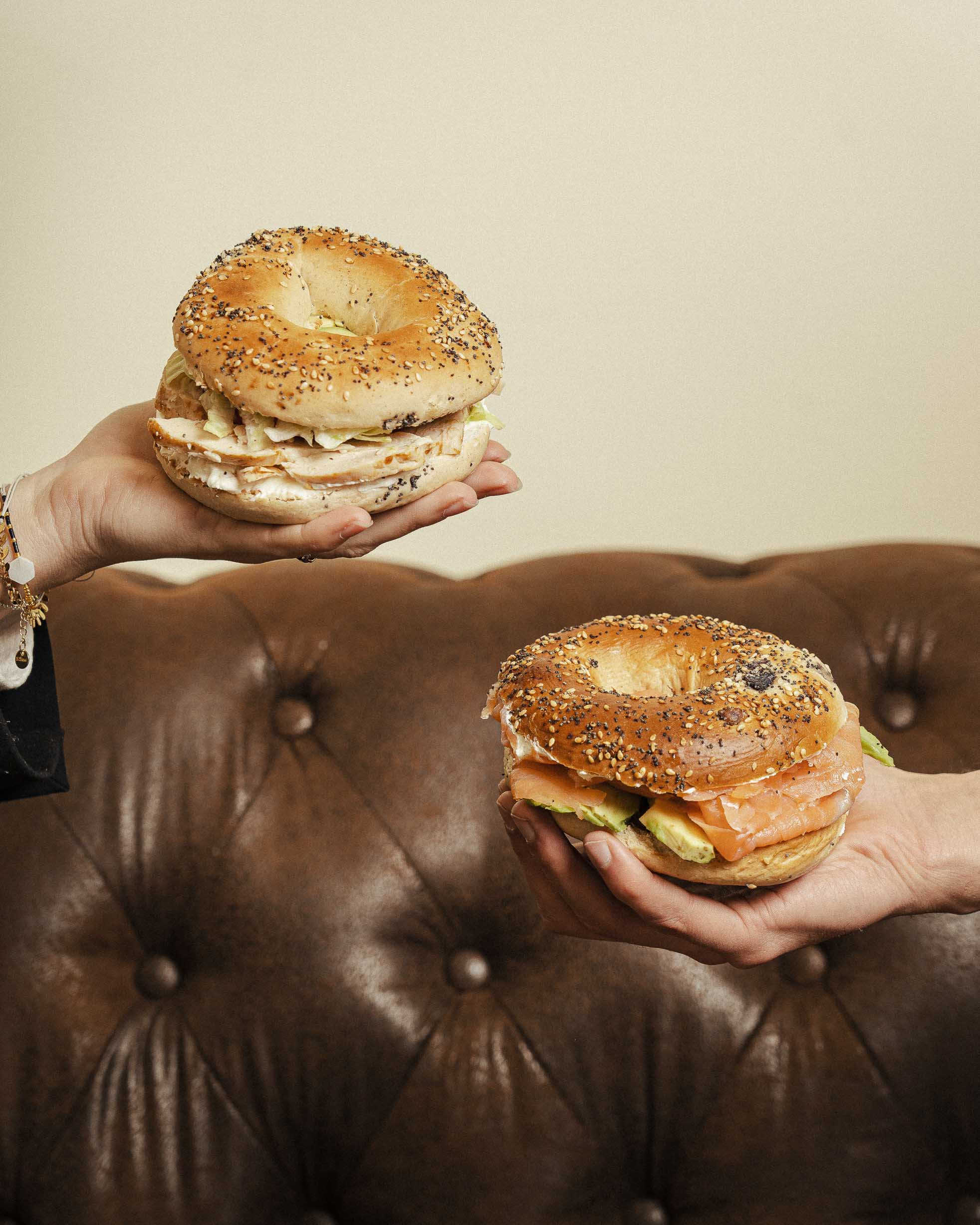 Deux bagels tenus à bout de bras devant un canapé en cuir. Shooting photo promotionnel pour Green Bagel Café à Chambéry.
