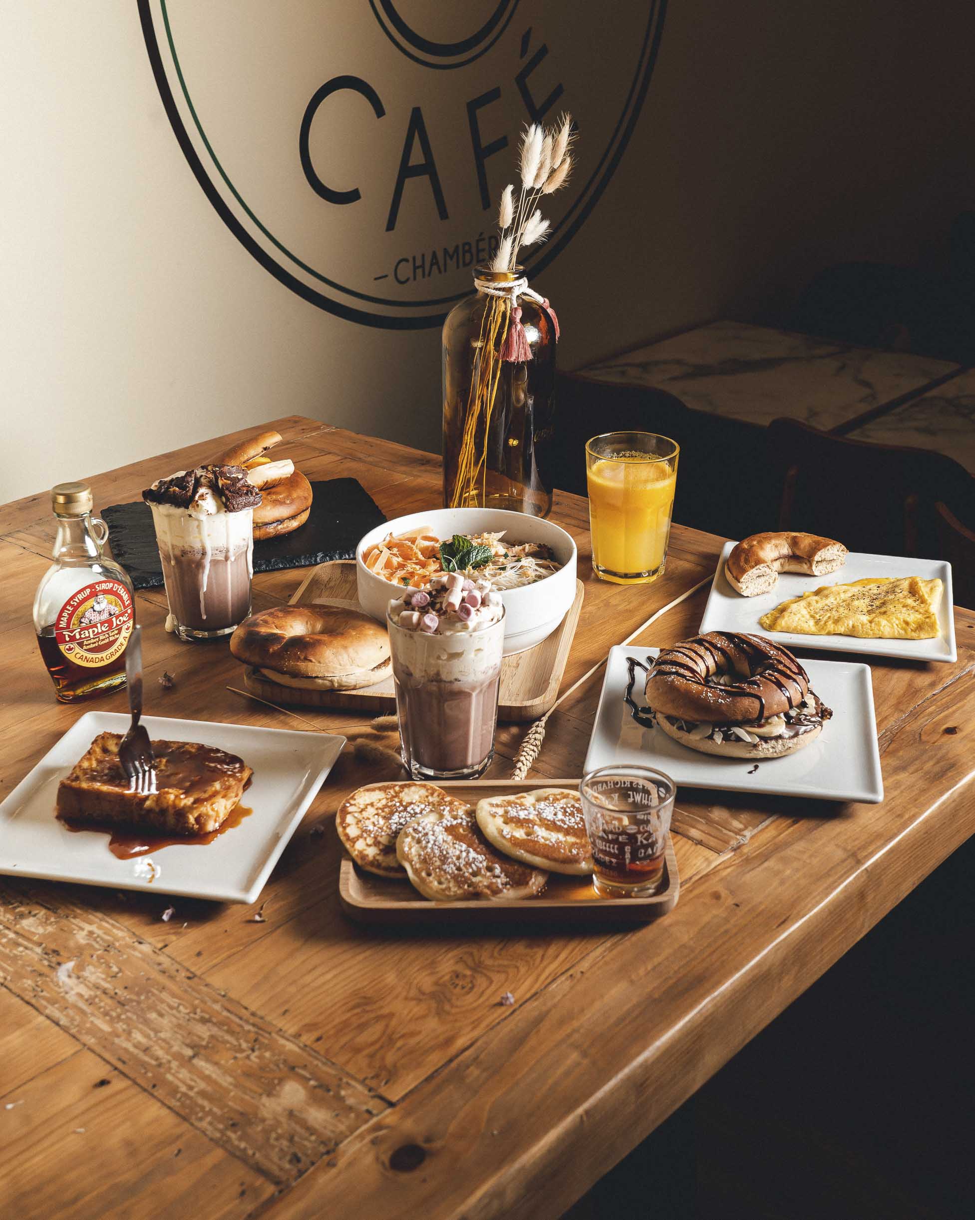 Un brunch : pancakes, bagels, pain perdu, omelette, chocolat et jus d'orange sur une table en bois. Shooting photo promotionnel pour Green Bagel Café à Chambéry.