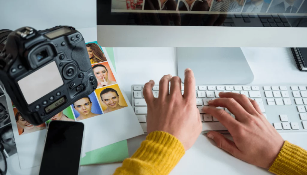 Photographe professionnel en train de retoucher des photos sur ordinateur, avec appareil photo reflex, smartphone et portraits imprimés, illustrant la différence entre photographie professionnelle et photo smartphone pour la communication d’entreprise.